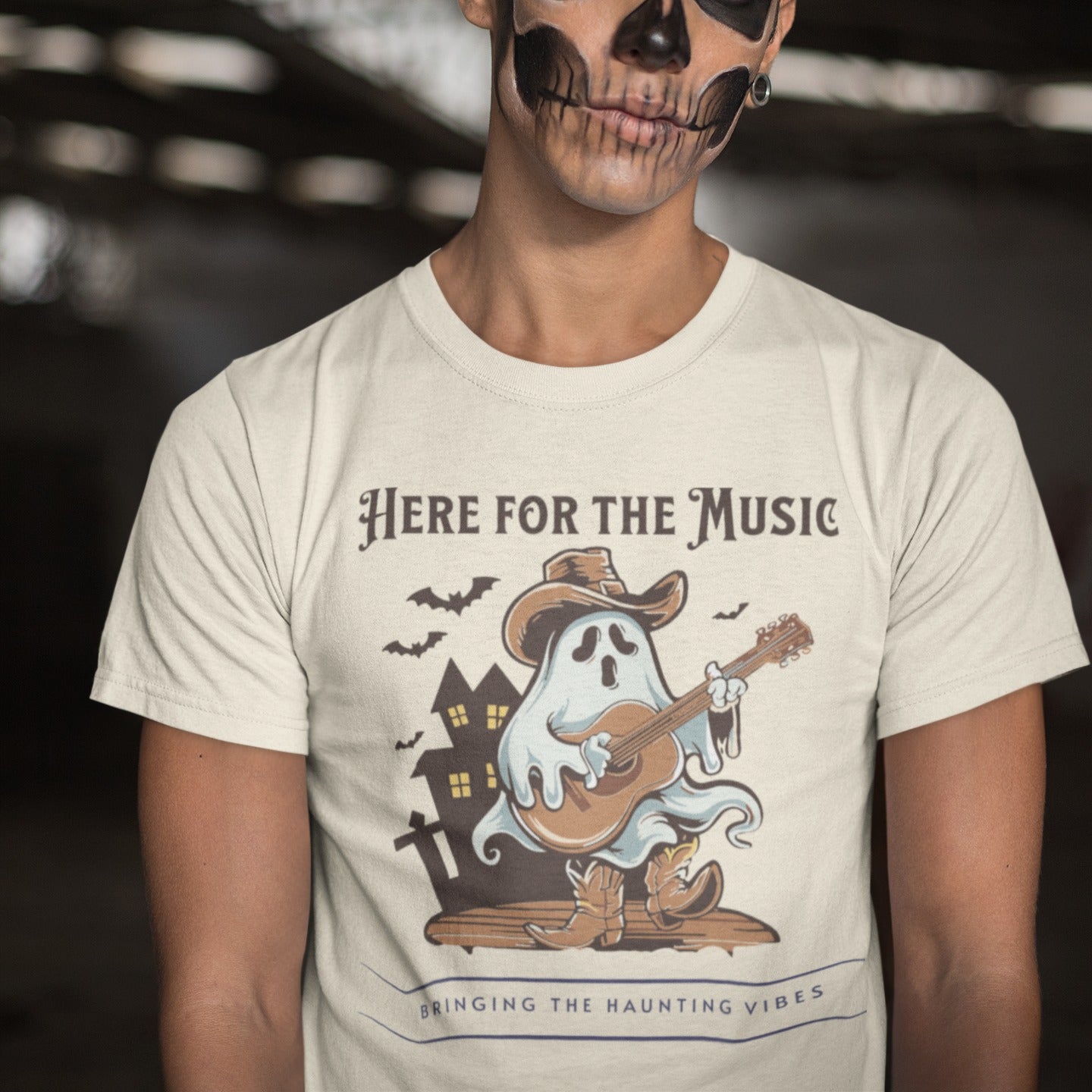 A light grey background features a stylized illustration of a white ghost wearing a brown cowboy hat and cowboy boots. The ghost is playing a brown acoustic guitar. Behind the figure, dark silhouettes of a haunted house and several bats are visible. At the top, the text reads "HERE FOR THE MUSIC" and at the bottom, "BRINGING THE HAUNTING VIBES". The design blends Halloween, music, and western themes.