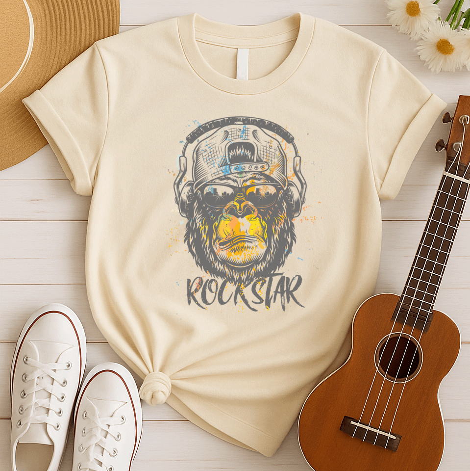 A grunge-style graphic features a detailed gorilla head wearing headphones, with the lower half of its face replaced by a textured splash of orange and yellow paint. The gorilla's fur and the headphones are rendered in shades of grey with distressed effects. Below the powerful primate, the word "ROCKSTAR" is emblazoned in a distressed white font with flecks of colorful paint matching the face. This striking design embodies raw power and musical rebellion.