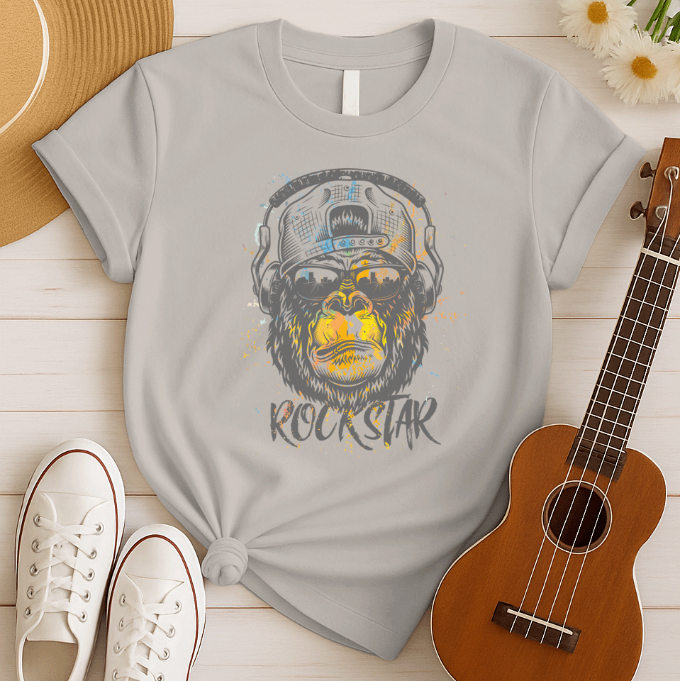 A grunge-style graphic features a detailed gorilla head wearing headphones, with the lower half of its face replaced by a textured splash of orange and yellow paint. The gorilla's fur and the headphones are rendered in shades of grey with distressed effects. Below the powerful primate, the word "ROCKSTAR" is emblazoned in a distressed white font with flecks of colorful paint matching the face. This striking design embodies raw power and musical rebellion.