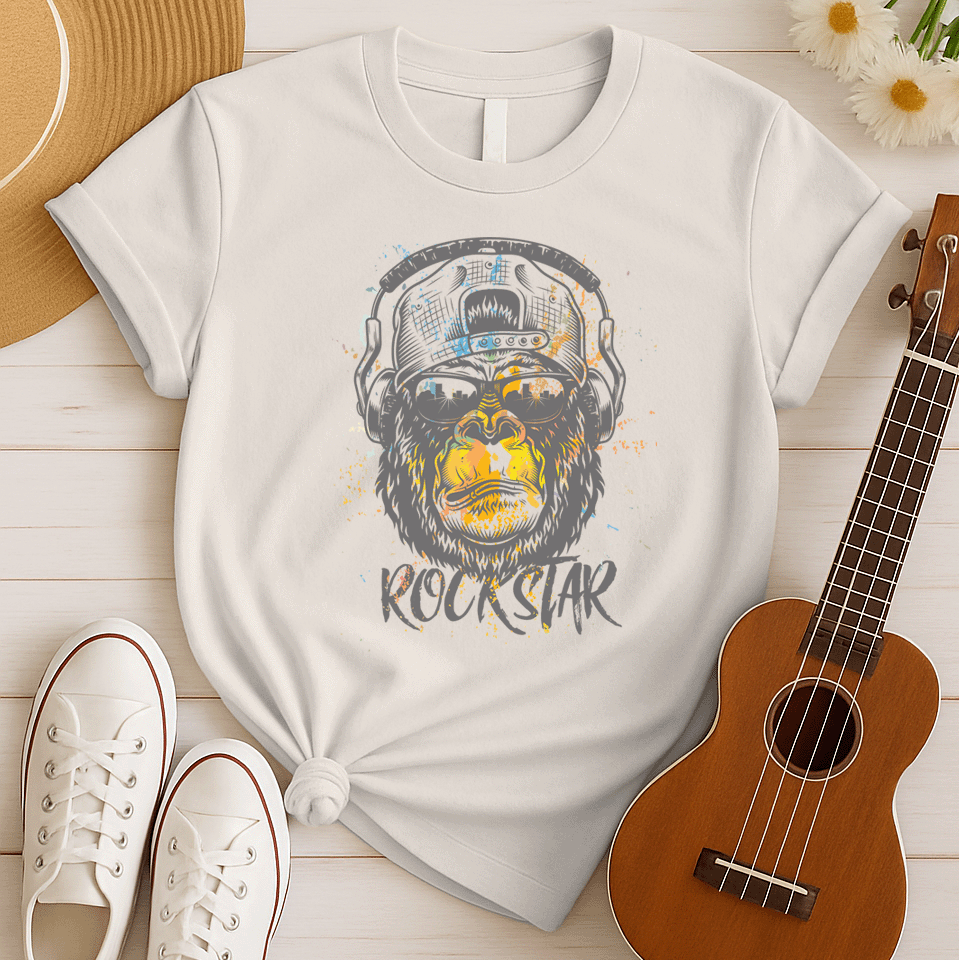 A grunge-style graphic features a detailed gorilla head wearing headphones, with the lower half of its face replaced by a textured splash of orange and yellow paint. The gorilla's fur and the headphones are rendered in shades of grey with distressed effects. Below the powerful primate, the word "ROCKSTAR" is emblazoned in a distressed white font with flecks of colorful paint matching the face. This striking design embodies raw power and musical rebellion.