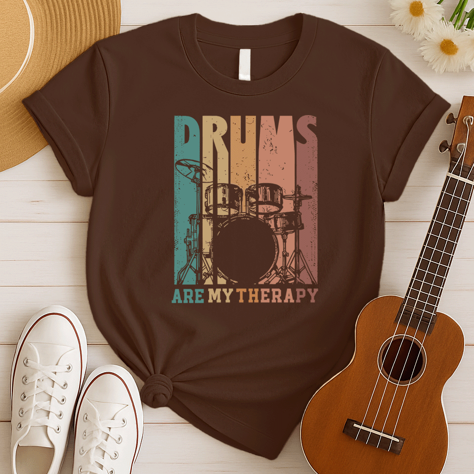 A black background features a stylized drum set (including bass drum, snare, toms, and cymbals) outlined in black. The drum set is filled with distressed vertical stripes of teal, brown, and orange/red. Above the drum set, the text "DRUMS" is in a large, distressed font with similar striped coloring. Below the drum set, the text "ARE MY THERAPY" is prominently displayed in orange. The design expresses a love for drumming, music, and percussion, ideal for drummers and music lovers.
