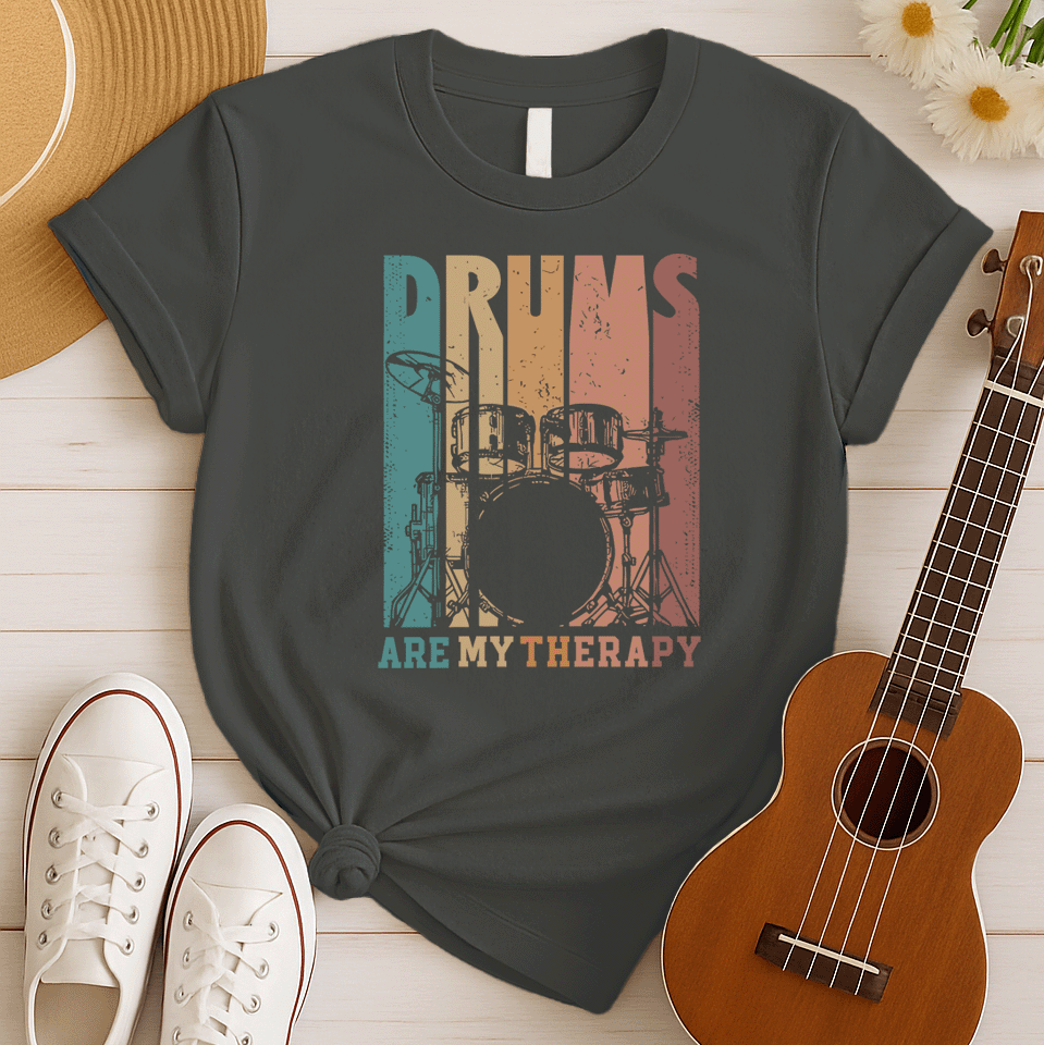 A black background features a stylized drum set (including bass drum, snare, toms, and cymbals) outlined in black. The drum set is filled with distressed vertical stripes of teal, brown, and orange/red. Above the drum set, the text "DRUMS" is in a large, distressed font with similar striped coloring. Below the drum set, the text "ARE MY THERAPY" is prominently displayed in orange. The design expresses a love for drumming, music, and percussion, ideal for drummers and music lovers.