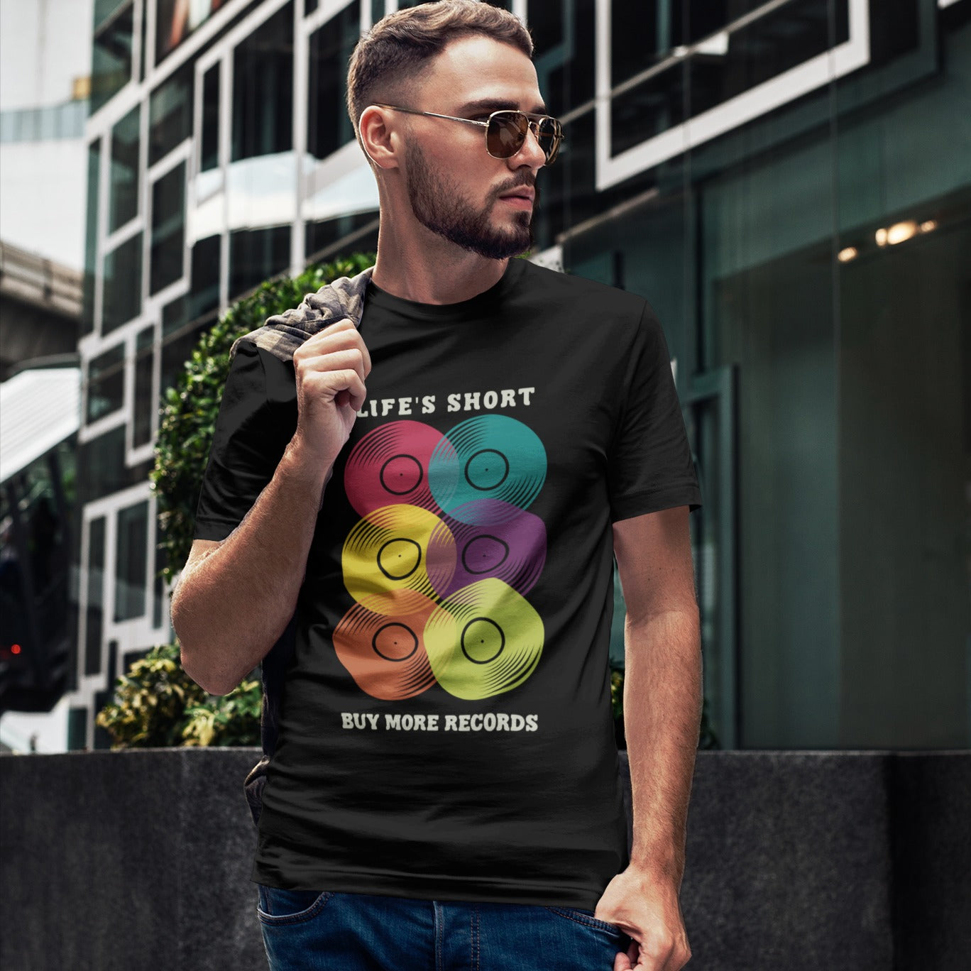 A black background features six vertically stacked, colorful vinyl records (LP records) with visible grooves and black center labels. The record colors include red, teal, yellow, purple, orange, and green. At the top, the distressed text "LIFE'S SHORT" is present, and at the bottom, the distressed text "BUY MORE RECORDS" is visible. The design is a clear celebration of vinyl culture and music collecting.