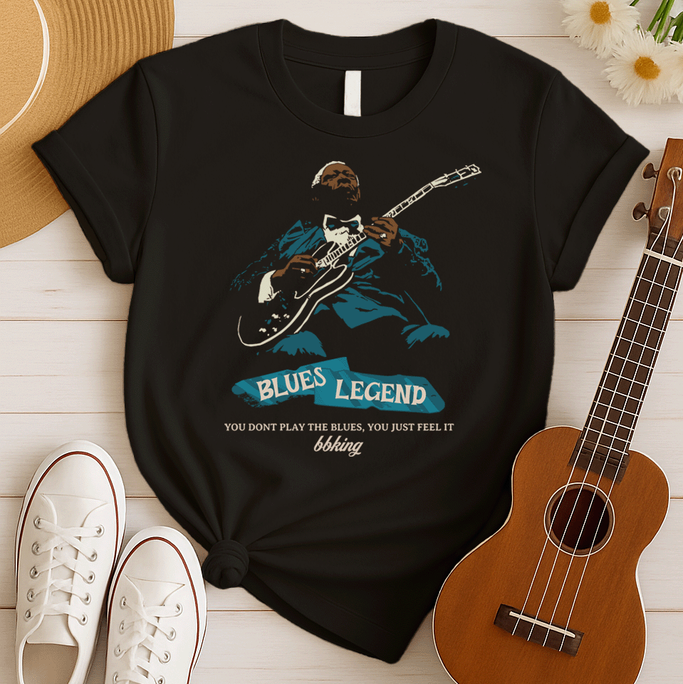 A black background features a stylized, silhouette-like illustration of a dark-skinned man in a suit and bow tie, playing an electric guitar. Highlights on his face, hands, guitar, and suit are in light brown/white and teal. Below the figure, two distressed, light blue shapes contain the text "BLUES LEGEND". Further below, the distressed white text reads "YOU DONT PLAY THE BLUES, YOU JUST FEEL IT", followed by "bbking" in a distressed, handwritten-style font. The design is a powerful tribute to blues music
