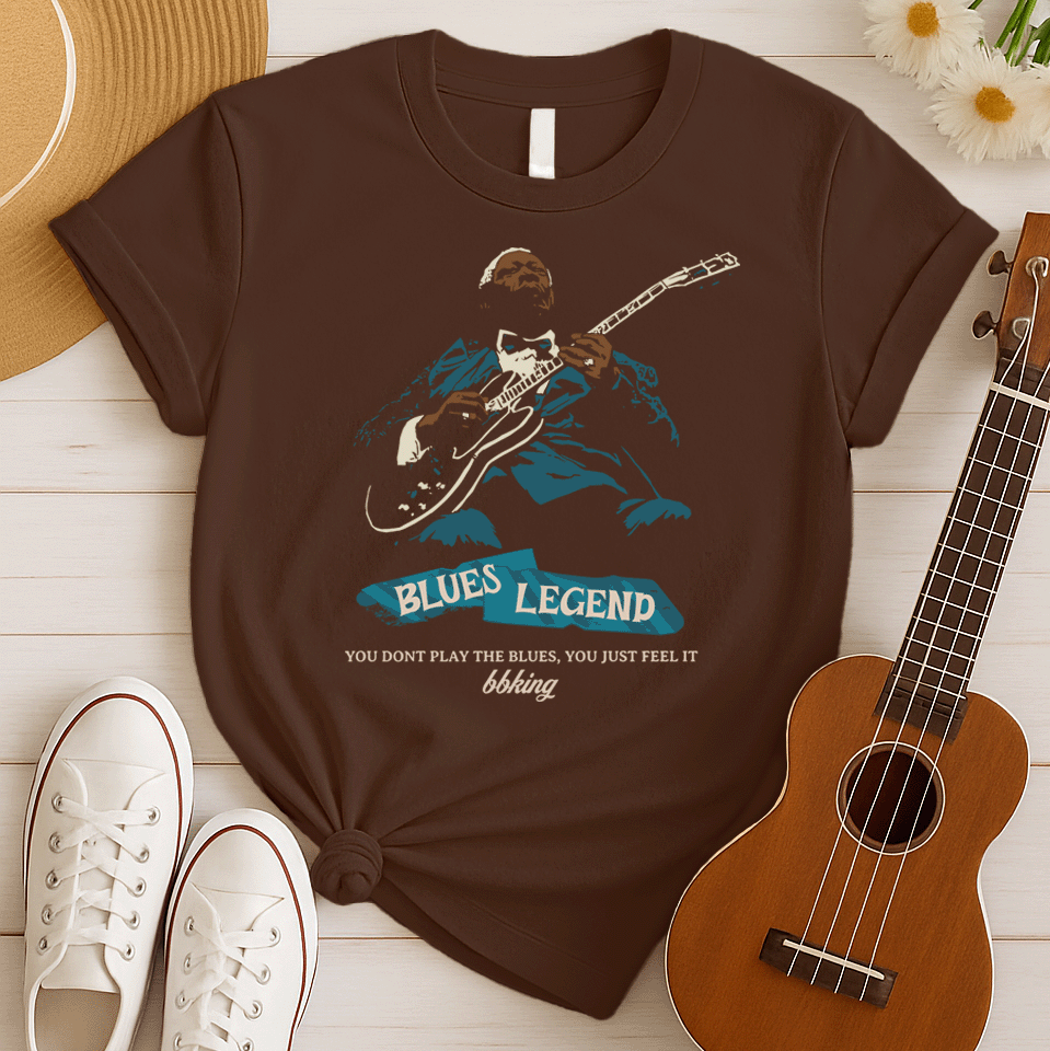 A black background features a stylized, silhouette-like illustration of a dark-skinned man in a suit and bow tie, playing an electric guitar. Highlights on his face, hands, guitar, and suit are in light brown/white and teal. Below the figure, two distressed, light blue shapes contain the text "BLUES LEGEND". Further below, the distressed white text reads "YOU DONT PLAY THE BLUES, YOU JUST FEEL IT", followed by "bbking" in a distressed, handwritten-style font. The design is a powerful tribute to blues music
