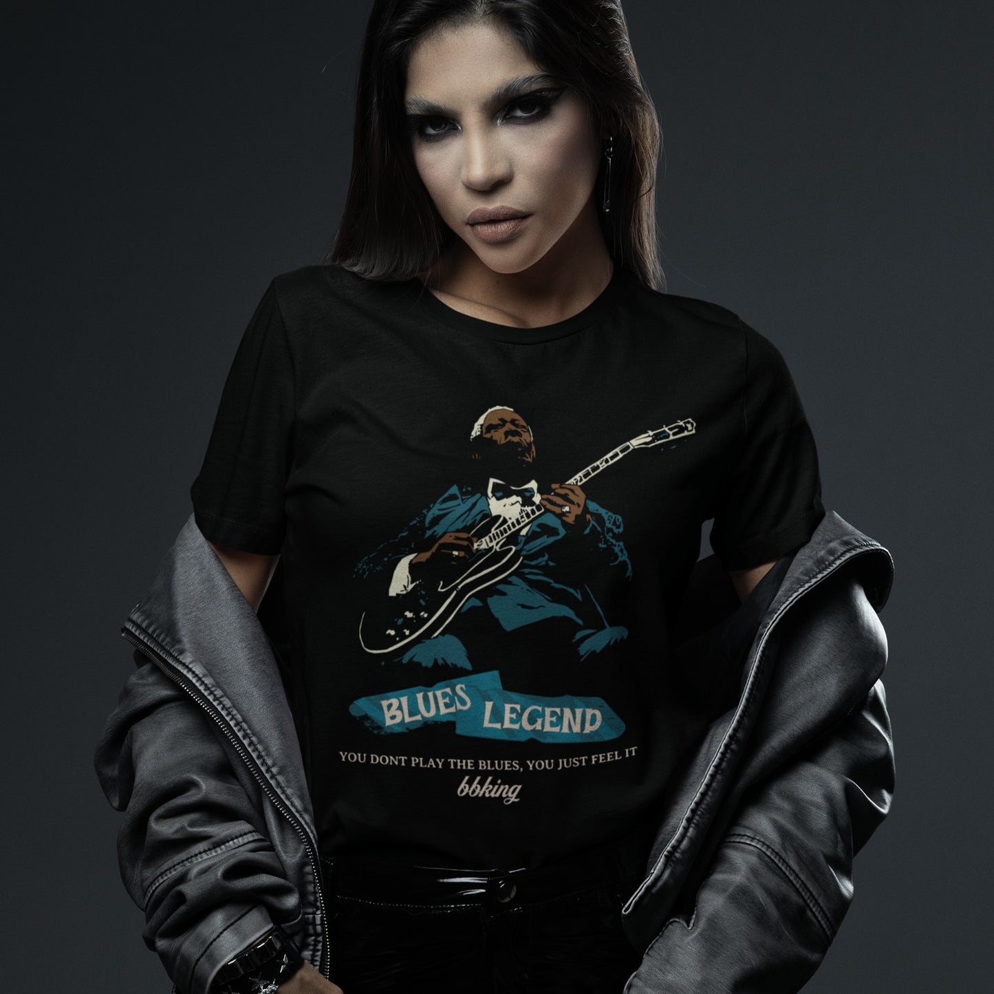 A black background features a stylized, silhouette-like illustration of a dark-skinned man in a suit and bow tie, playing an electric guitar. Highlights on his face, hands, guitar, and suit are in light brown/white and teal. Below the figure, two distressed, light blue shapes contain the text "BLUES LEGEND". Further below, the distressed white text reads "YOU DONT PLAY THE BLUES, YOU JUST FEEL IT", followed by "bbking" in a distressed, handwritten-style font. The design is a powerful tribute to blues music