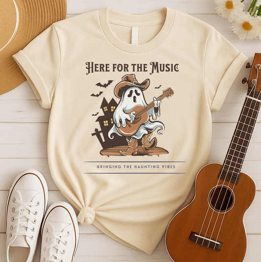 A light grey background features a stylized illustration of a white ghost wearing a brown cowboy hat and cowboy boots. The ghost is playing a brown acoustic guitar. Behind the figure, dark silhouettes of a haunted house and several bats are visible. At the top, the text reads "HERE FOR THE MUSIC" and at the bottom, "BRINGING THE HAUNTING VIBES". The design blends Halloween, music, and western themes.