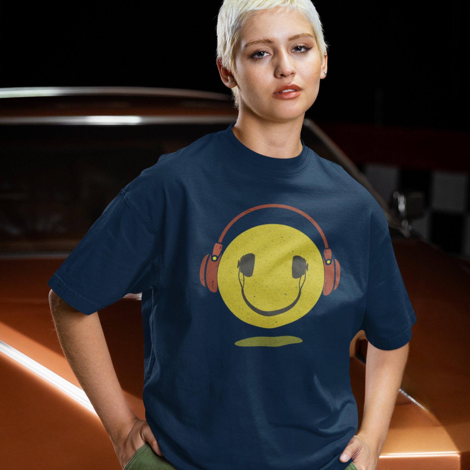 A bold and expressive graphic features a textured, distressed yellow smiley face with a prominent black smile and eyes. Large, rustic orange over-ear headphones, also with a distressed texture, are worn on top of the smiley face. The entire design creates a lively and vintage-inspired image that conveys happiness and a love for music.