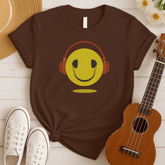 A bold and expressive graphic features a textured, distressed yellow smiley face with a prominent black smile and eyes. Large, rustic orange over-ear headphones, also with a distressed texture, are worn on top of the smiley face. The entire design creates a lively and vintage-inspired image that conveys happiness and a love for music.