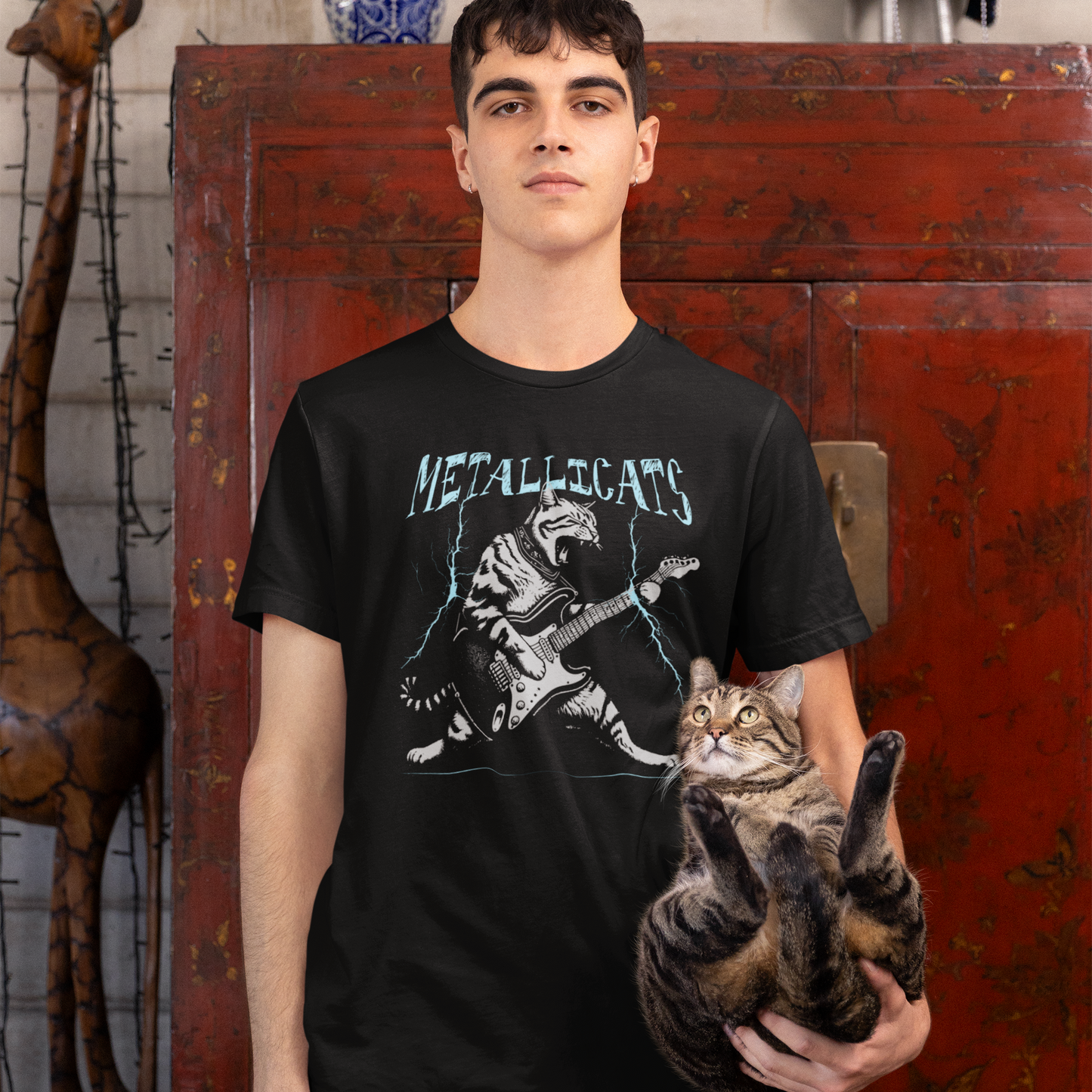 Dynamic graphic of a gray striped tabby cat with its mouth open, appearing to roar, passionately playing a black and white electric guitar. Blue lightning bolts emanate from the guitar. Text above reads "METALLICATS". The cat wears a studded collar. Ideal for heavy metal fans, rock music lovers, guitarists, and cat lovers.