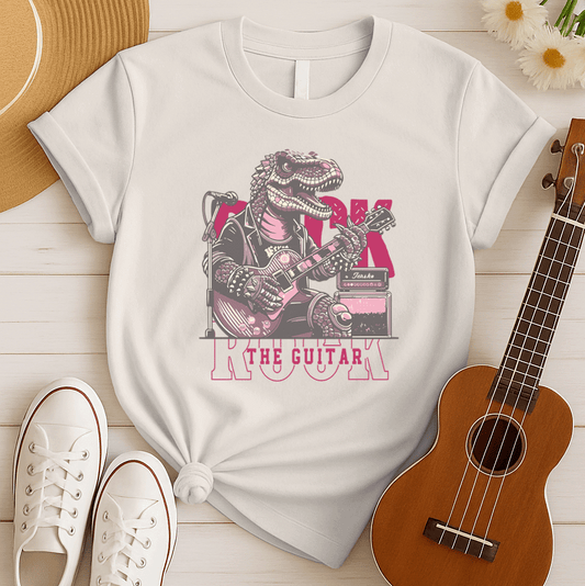 Dynamic graphic of a large, muscular pink/purple T-Rex dinosaur wearing a black studded leather jacket, intensely playing a pink electric guitar connected to a guitar amplifier and microphone. Large pink distressed text behind the T-Rex reads "ROCK" and bold pink text below reads "THE GUITAR". The design embodies rock music, dinosaur power, and guitar shredding, ideal for rock music fans, guitarists, and dinosaur enthusiasts.
