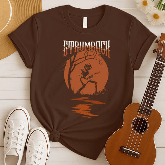 Stylized graphic of a silhouetted guitarist in a hat and jacket, passionately playing an acoustic or electric guitar against a large, glowing orange full moon. A bare tree branch extends over them. Orange text above reads "STRUMROCK". The scene evokes a rock concert or solo performance under the night sky, ideal for guitar players, rock music fans, and musicians.