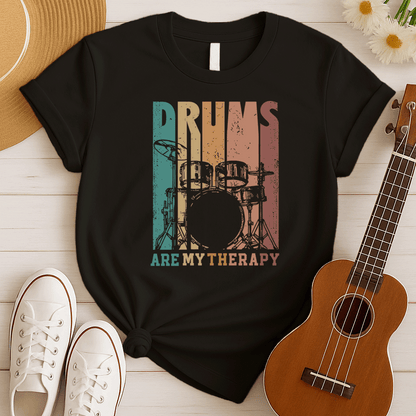 A black background features a stylized drum set (including bass drum, snare, toms, and cymbals) outlined in black. The drum set is filled with distressed vertical stripes of teal, brown, and orange/red. Above the drum set, the text "DRUMS" is in a large, distressed font with similar striped coloring. Below the drum set, the text "ARE MY THERAPY" is prominently displayed in orange. The design expresses a love for drumming, music, and percussion, ideal for drummers and music lovers.