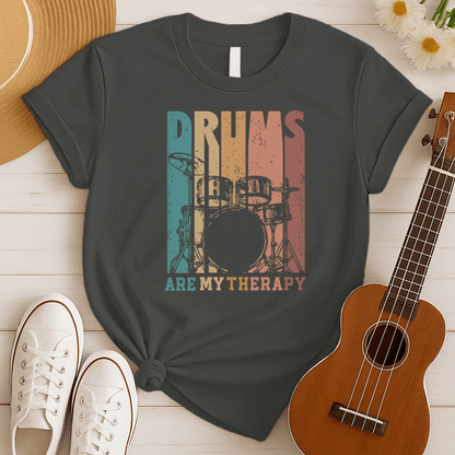 A black background features a stylized drum set (including bass drum, snare, toms, and cymbals) outlined in black. The drum set is filled with distressed vertical stripes of teal, brown, and orange/red. Above the drum set, the text "DRUMS" is in a large, distressed font with similar striped coloring. Below the drum set, the text "ARE MY THERAPY" is prominently displayed in orange. The design expresses a love for drumming, music, and percussion, ideal for drummers and music lovers.