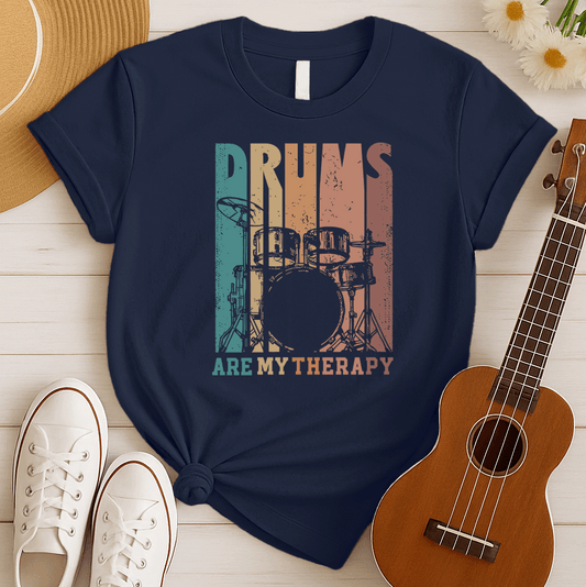 A black background features a stylized drum set (including bass drum, snare, toms, and cymbals) outlined in black. The drum set is filled with distressed vertical stripes of teal, brown, and orange/red. Above the drum set, the text "DRUMS" is in a large, distressed font with similar striped coloring. Below the drum set, the text "ARE MY THERAPY" is prominently displayed in orange. The design expresses a love for drumming, music, and percussion, ideal for drummers and music lovers.