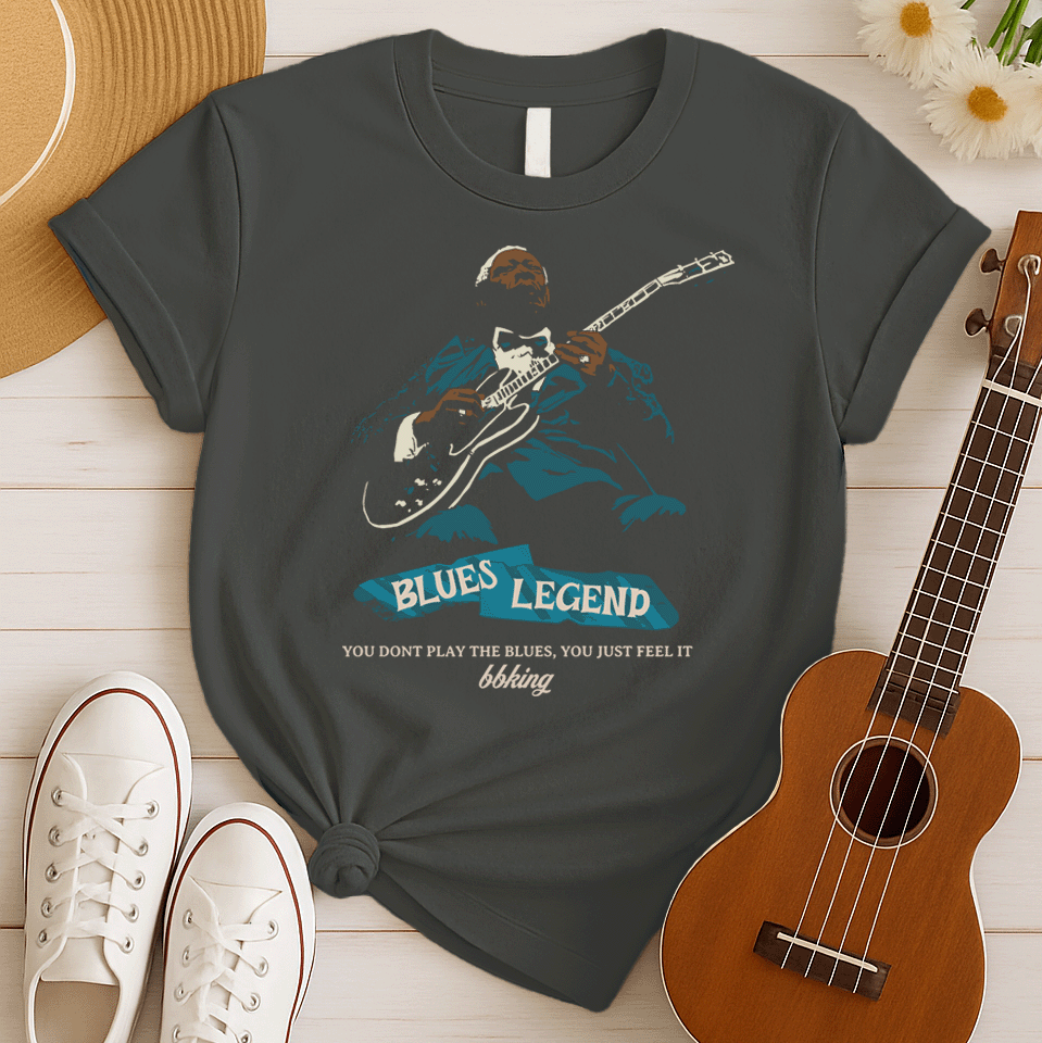 A black background features a stylized, silhouette-like illustration of a dark-skinned man in a suit and bow tie, playing an electric guitar. Highlights on his face, hands, guitar, and suit are in light brown/white and teal. Below the figure, two distressed, light blue shapes contain the text "BLUES LEGEND". Further below, the distressed white text reads "YOU DONT PLAY THE BLUES, YOU JUST FEEL IT", followed by "bbking" in a distressed, handwritten-style font. The design is a powerful tribute to blues music 