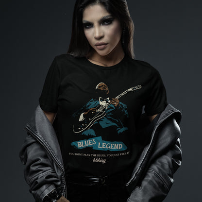 A black background features a stylized, silhouette-like illustration of a dark-skinned man in a suit and bow tie, playing an electric guitar. Highlights on his face, hands, guitar, and suit are in light brown/white and teal. Below the figure, two distressed, light blue shapes contain the text "BLUES LEGEND". Further below, the distressed white text reads "YOU DONT PLAY THE BLUES, YOU JUST FEEL IT", followed by "bbking" in a distressed, handwritten-style font. The design is a powerful tribute to blues music 
